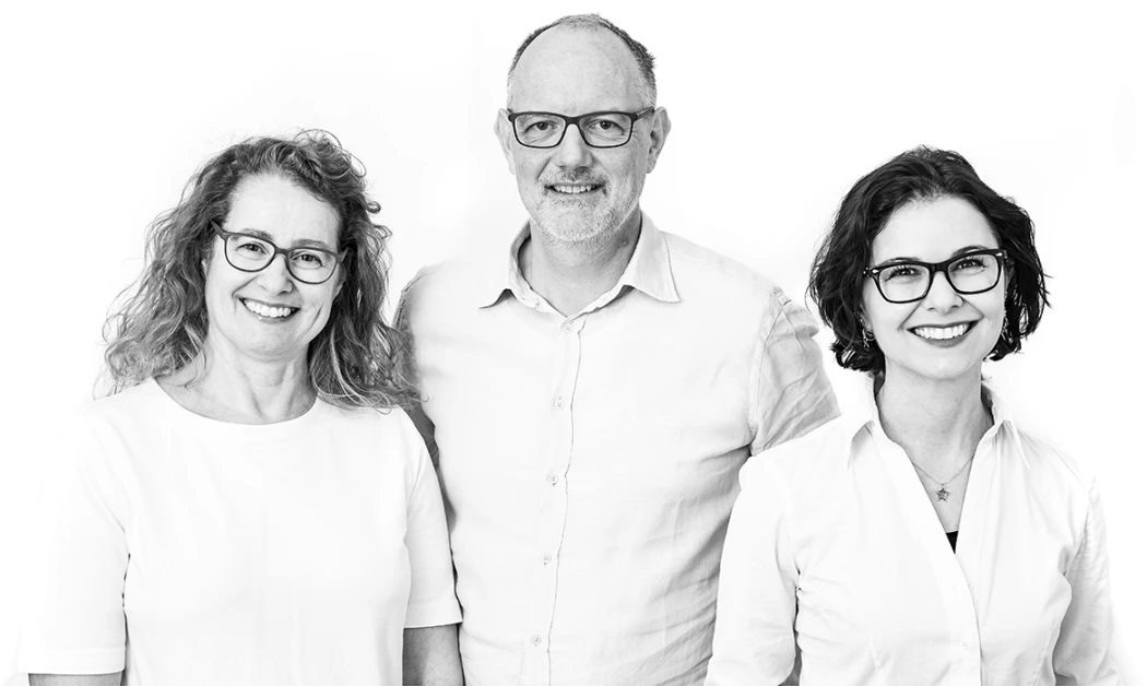Portraitfoto von drei freundlich laechelnden Teammitgliedern – zwei Frauen und ein Mann – in heller Kleidung, nebeneinander stehend