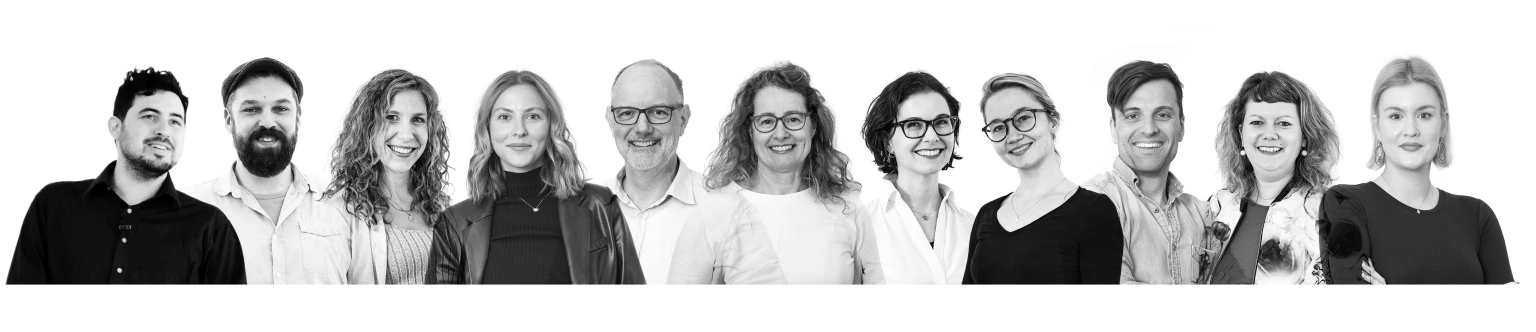 Schwarz-weiß-Gruppenfoto von 11 fröhlichen Menschen unterschiedlichen Alters, die nebeneinander stehen und in die Kamera lächeln.