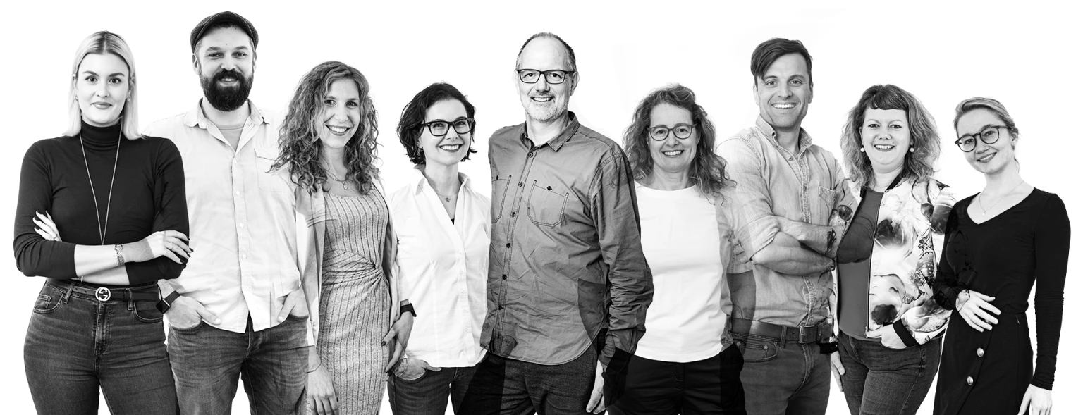 Schwarz-weiß-Gruppenfoto von 11 fröhlichen Menschen unterschiedlichen Alters, die nebeneinander stehen und in die Kamera lächeln.