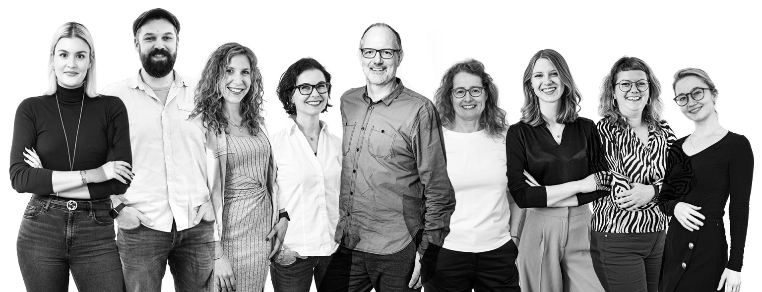 Schwarz-weiß-Gruppenfoto von 11 fröhlichen Menschen unterschiedlichen Alters, die nebeneinander stehen und in die Kamera lächeln.