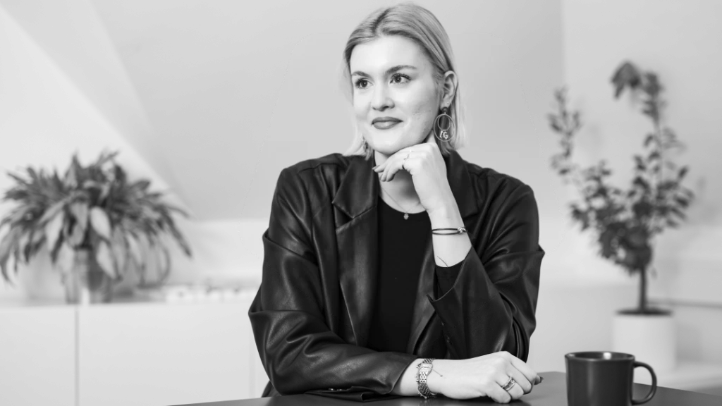 Junge Frau mit hellem, schulterlangem Haar, auffälligen Ohrringen und Lederjacke blickt nachdenklich zur Seite, die Hand ans Kinn gelegt.