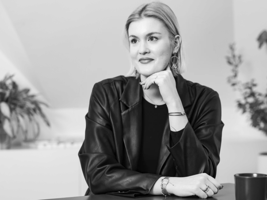 Junge Frau mit hellem, schulterlangem Haar, auffälligen Ohrringen und Lederjacke blickt nachdenklich zur Seite, die Hand ans Kinn gelegt.