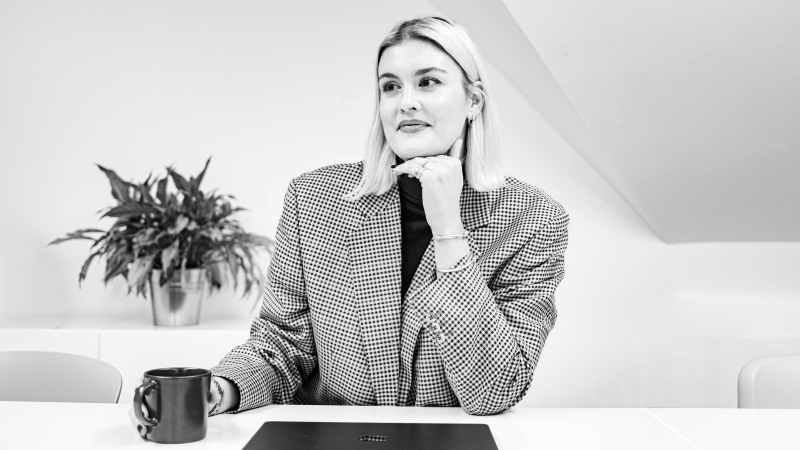 Junge Frau mit hellem, schulterlangem Haar, auffälligen Ohrringen und Lederjacke blickt nachdenklich zur Seite, die Hand ans Kinn gelegt.