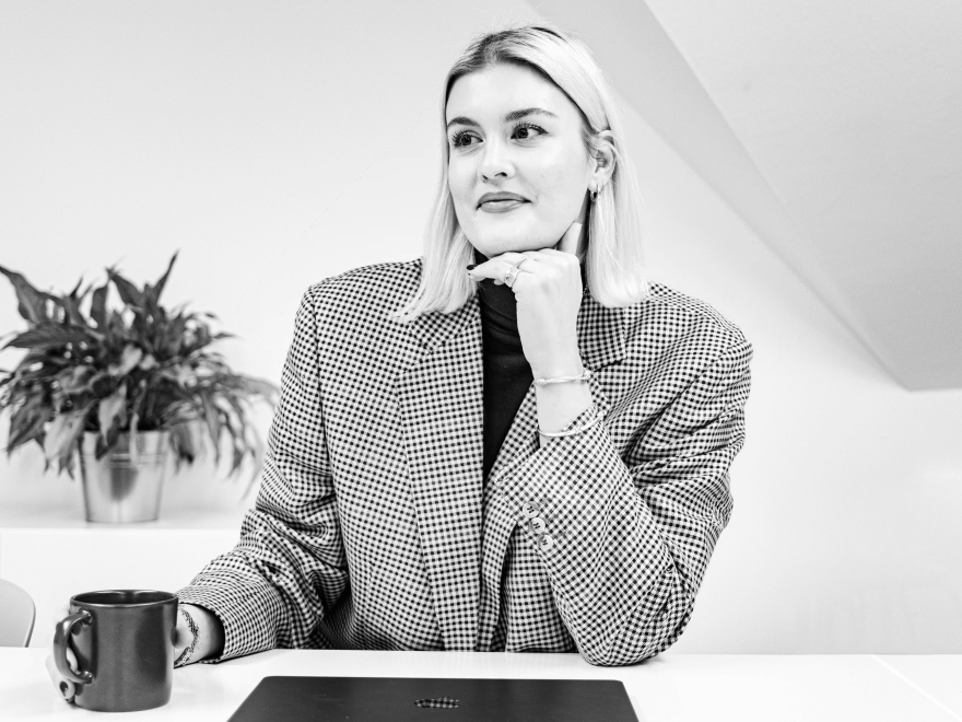 Junge Frau mit hellem, schulterlangem Haar, auffälligen Ohrringen und Lederjacke blickt nachdenklich zur Seite, die Hand ans Kinn gelegt.