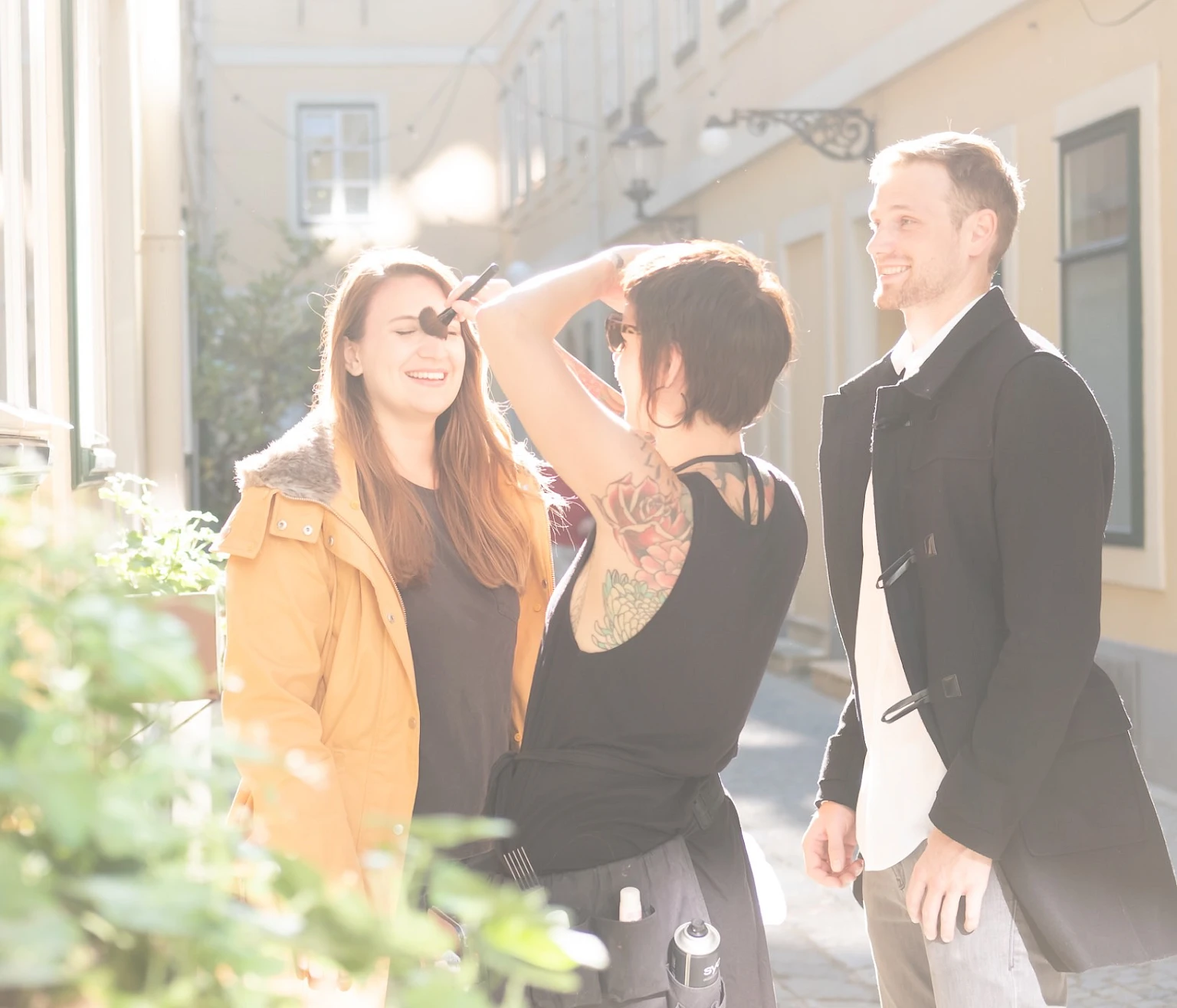 Making-of-Szene bei einem Fotoshooting im Freien: Visagistin pudert das Gesicht einer jungen Frau, daneben steht ein laechelnder Mann. Sonniger Innenhof mit heller Hausfassade im Hintergrund