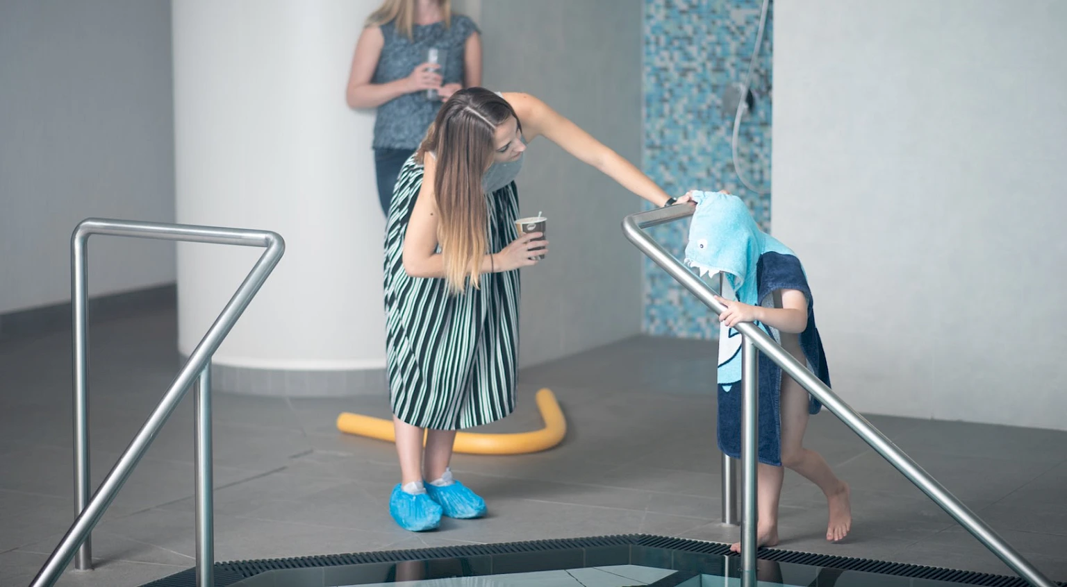 Making-of-Szene eines Shootings fuer die Awareness-Kampagne am Rand eines Indoor-Pools: Frau im Kleid und blauen Ueberschuhen betreut ein Kind im Hai-Badeponcho, das die Treppe ins Becken hinuntersteigt
