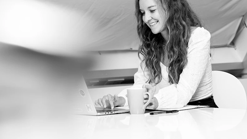 Laechelnde Frau sitzt mit Tasse bei Schreibtisch mit Laptop