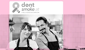 Zwei freundlich lächelnde Menschen in Kochkleidung stehen nebeneinander in einer Küche, darüber das Logo der Initiative dontsmoke.at Volksbegehren