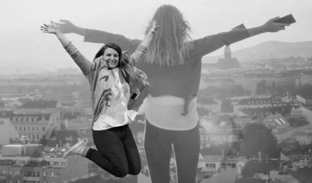 Eine Frau springt in die Luft mit ausgebreiteten Armen, im Hintergrund blickt eine transparente Silhouette einer anderen Person mit erhobenen Armen ueber eine Stadtlandschaft