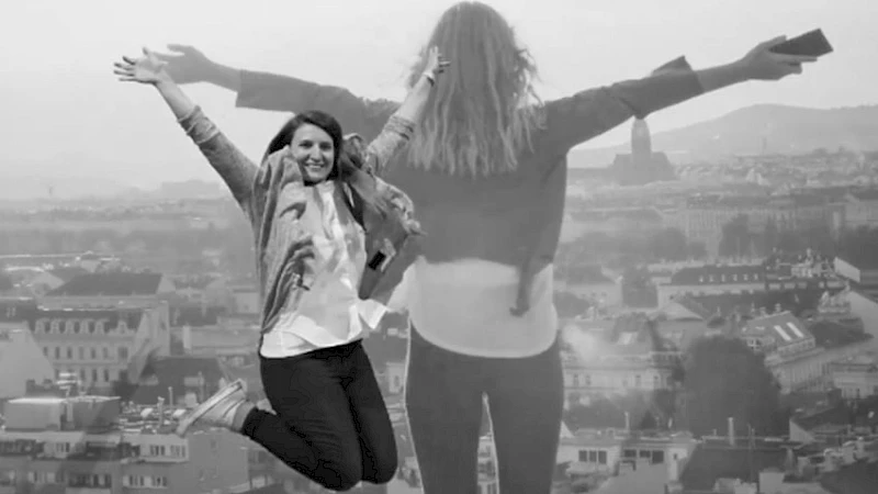 Eine Frau springt in die Luft mit ausgebreiteten Armen, im Hintergrund blickt eine transparente Silhouette einer anderen Person mit erhobenen Armen ueber eine Stadtlandschaft