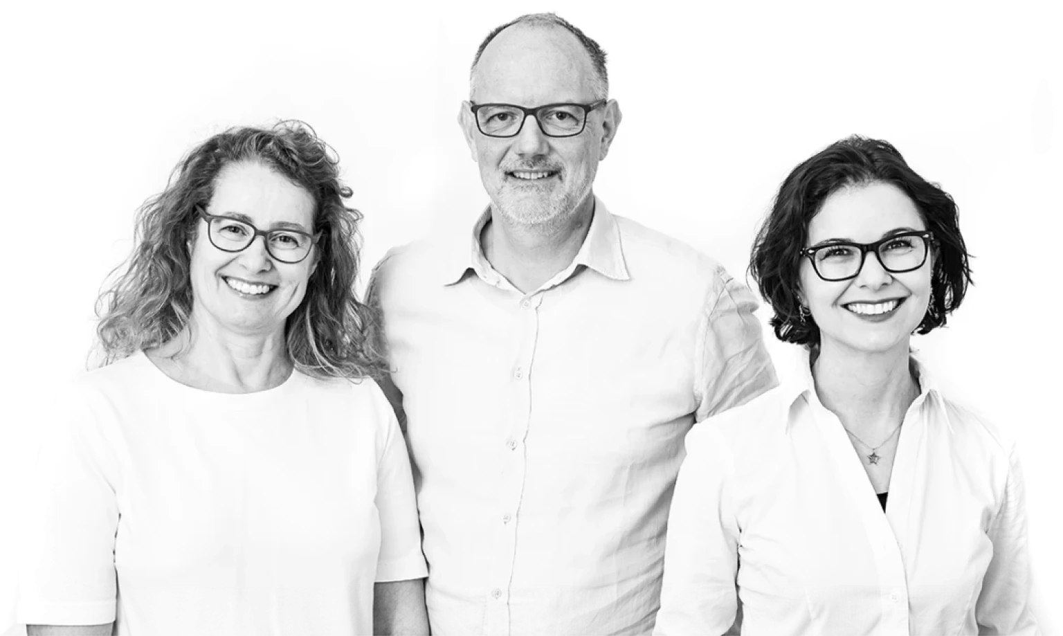 Schwarz-Weiß-Teamfoto der drei Führungspersönlichkeiten: Gründer, Gründerin und Partnerin lächeln gemeinsam in die Kamera. Alle tragen Brillen und helle Kleidung.