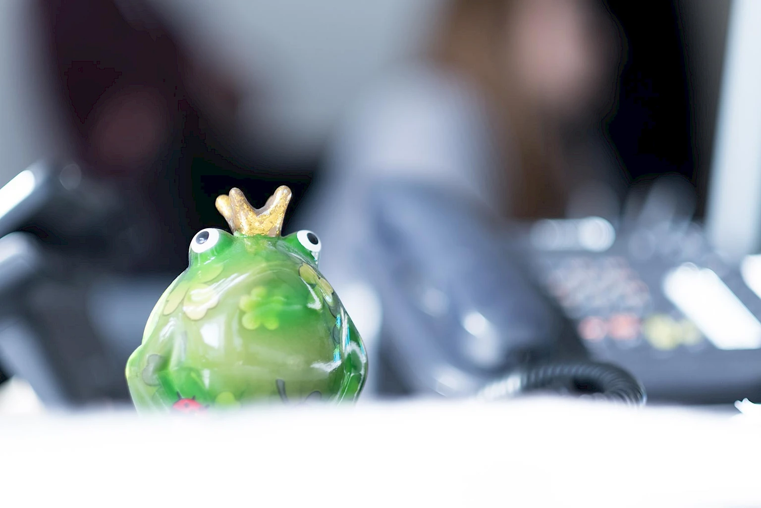 Froschspielzeug auf einem Schreibtisch mit Festnetz-Telefon und Menschen im Hintergrund