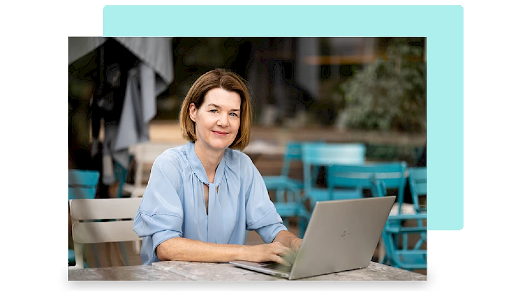 Freundliche Frau sitzt in einem Garten bei Tisch mit einem aufgeklappten Laptop und arbeitet.